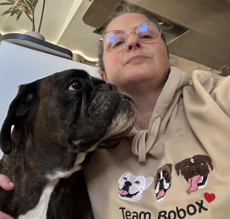 Une femme en sweat à capuche 'Team Boxer' avec des illustrations de chiens, serrant un boxer noir et blanc contre elle